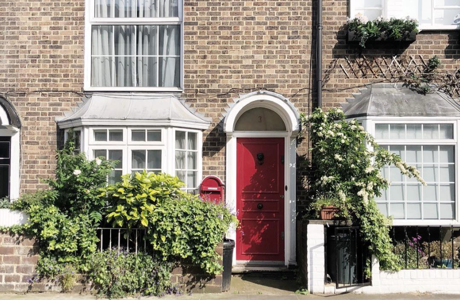 cropped brick house in london with white windows, some foliage, and a red door